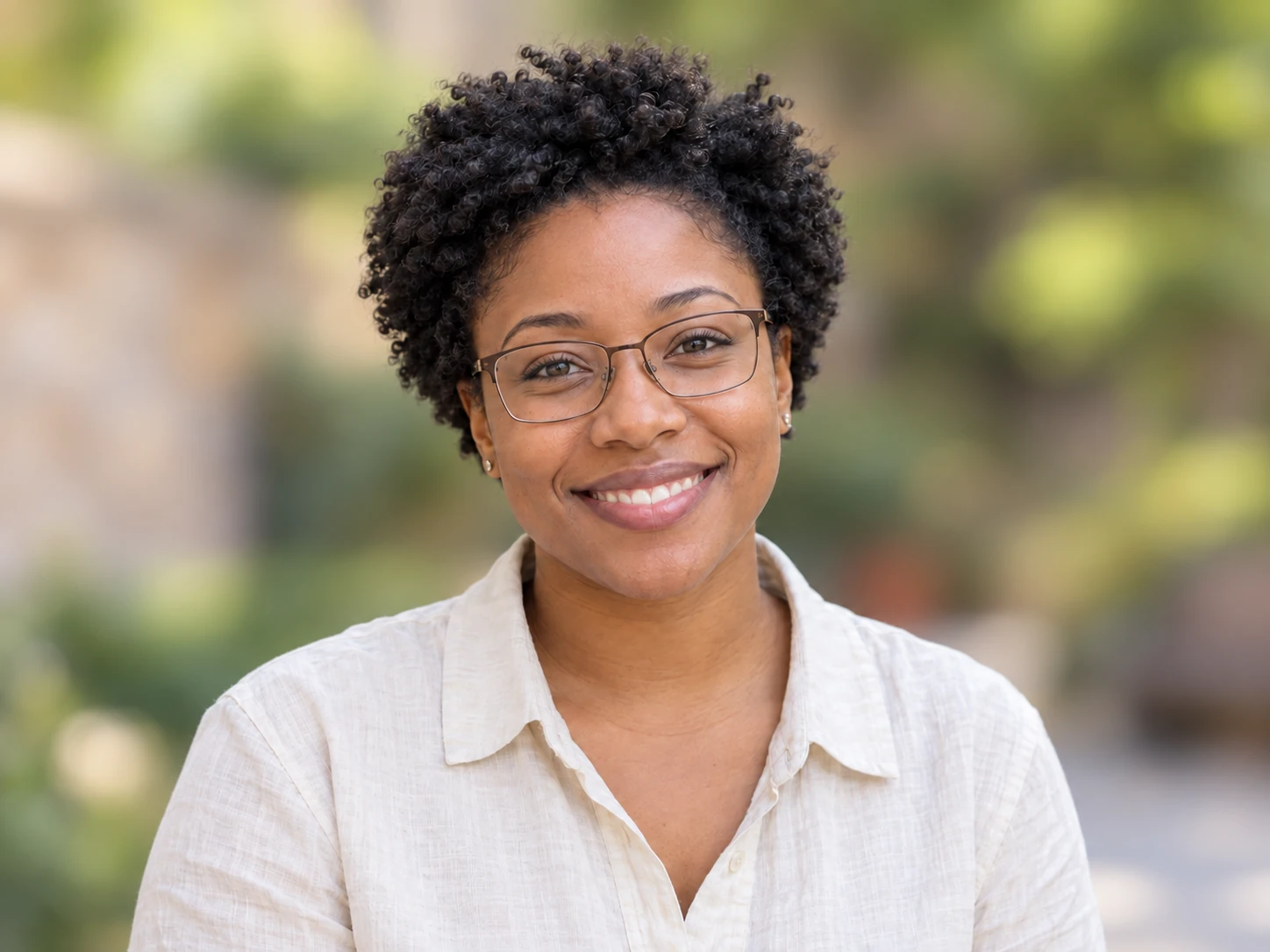 Retrato natural y sonriente de una mujer con el pelo corto y rizado, gafas rectangulares y camisa color crema frente a un patio muy desenfocado, encuadrada para una foto de perfil