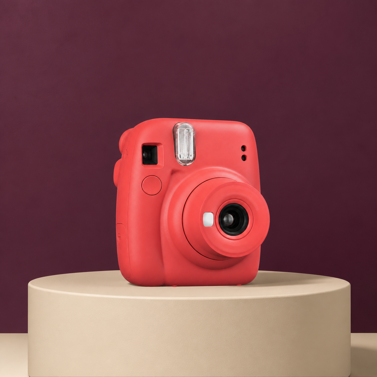 Coral instant camera on a warm-cream display pedestal against a plum background.